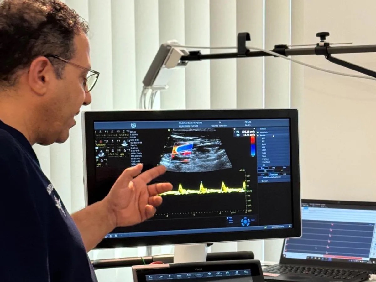 Dr. Qedra zeigt auf einen Ultraschall-Monitor, auf dem der Blutfluss in einer Arterie zu sehen ist.